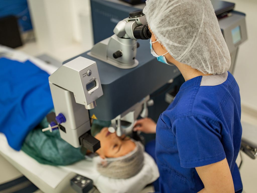 A healthcare professional in a blue scrub and surgical cap is operating a laser eye surgery machine on a patient lying down. The patient's eyes are covered with protective gear, and the professional is focused on adjusting the equipment in a well-lit surgical room