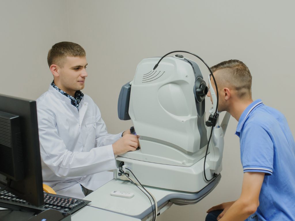 A young male optometrist in a white lab coat operates an optical coherence tomography machine, assisting a young male patient seated and peering into the device in a clinical setting.