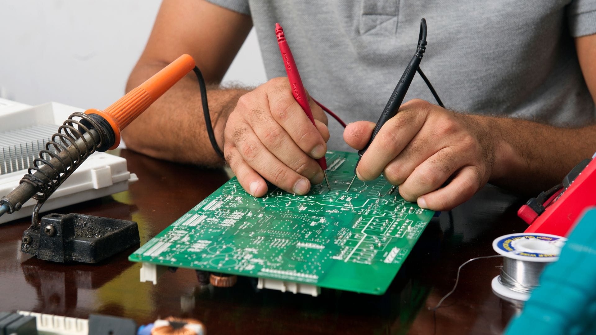 Person using multimeter probes to test or troubleshoot a green circuit board, with a soldering iron nearby.